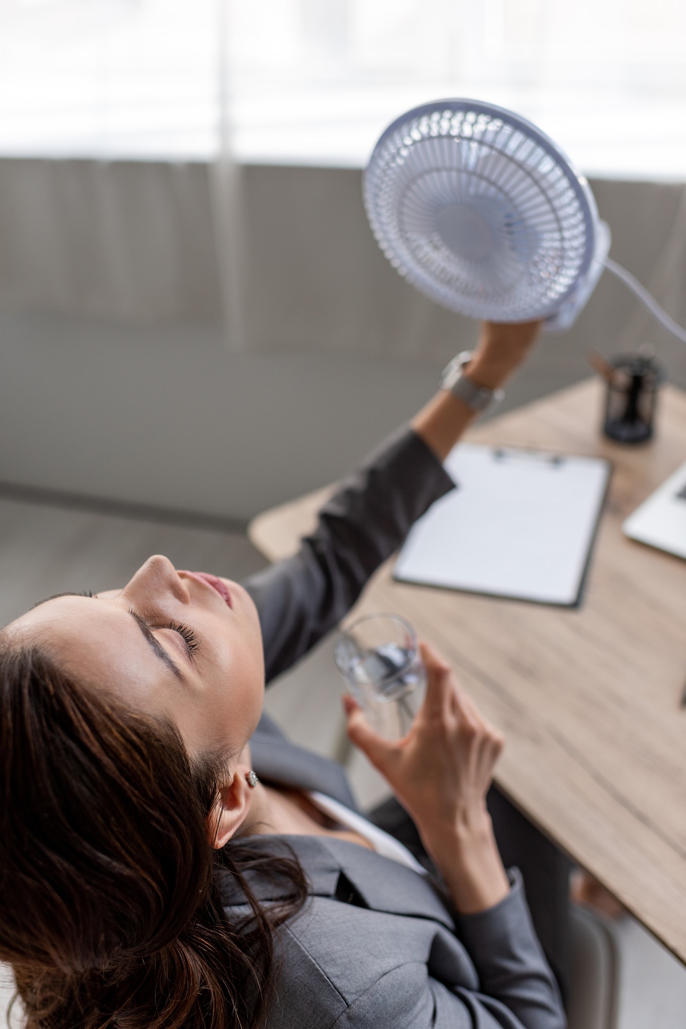 overhead view of pretty businesswoman holding blowing electric fan and glass of water while